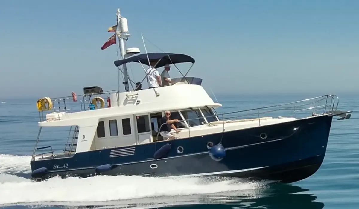 A cream and navy motor yacht moving through calm blue sea with crew members visible on deck under clear skies.