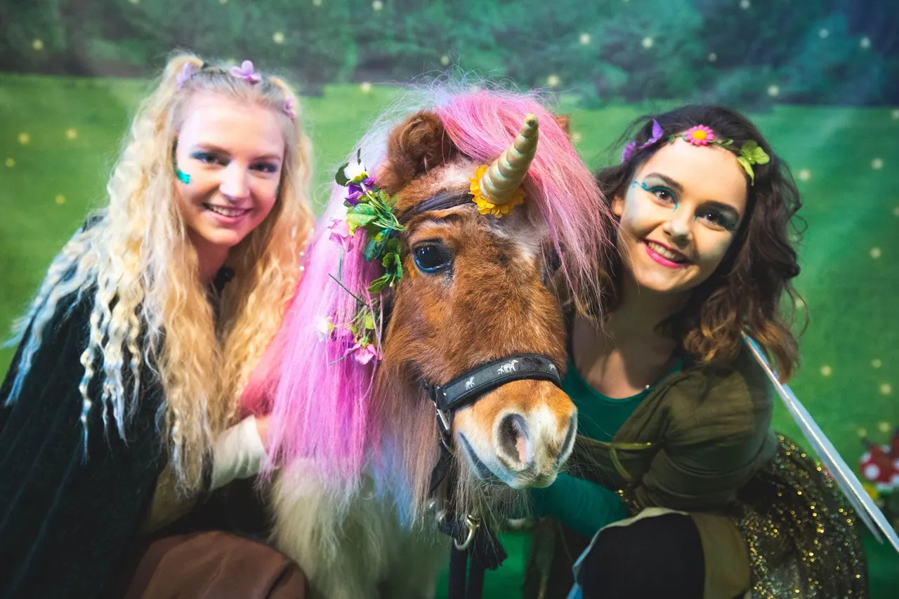 Three young people posing with a decorative pony dressed with pink hair and floral accessories in front of a green backdrop.