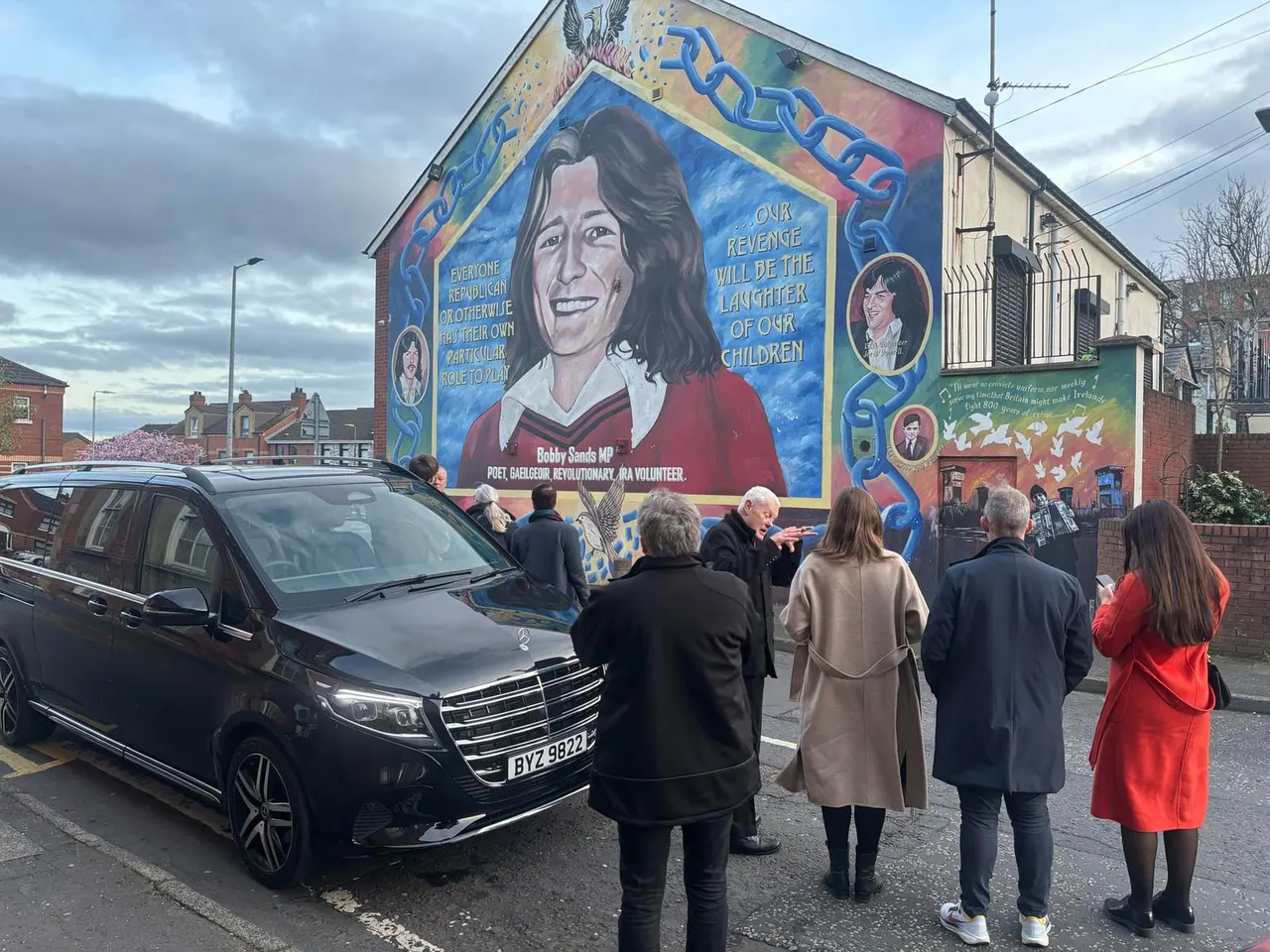 People gathered on a street viewing a large colourful mural on a brick building, with a black car parked nearby.