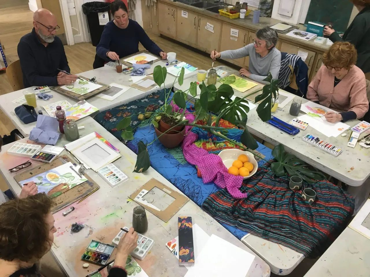 Adults painting with watercolours around tables in an art studio, with potted plants and fruit as still life subjects in the centre.