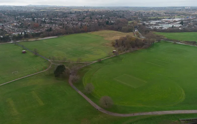 Aerial view of sports fields and green spaces with residential suburbs and distant hills beyond.