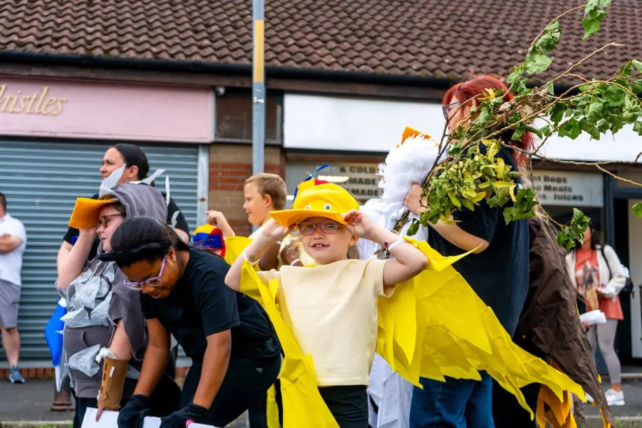 Children in fancy dress, including one wearing a yellow superhero outfit and hat, participating in a street parade or outdoor performance event.