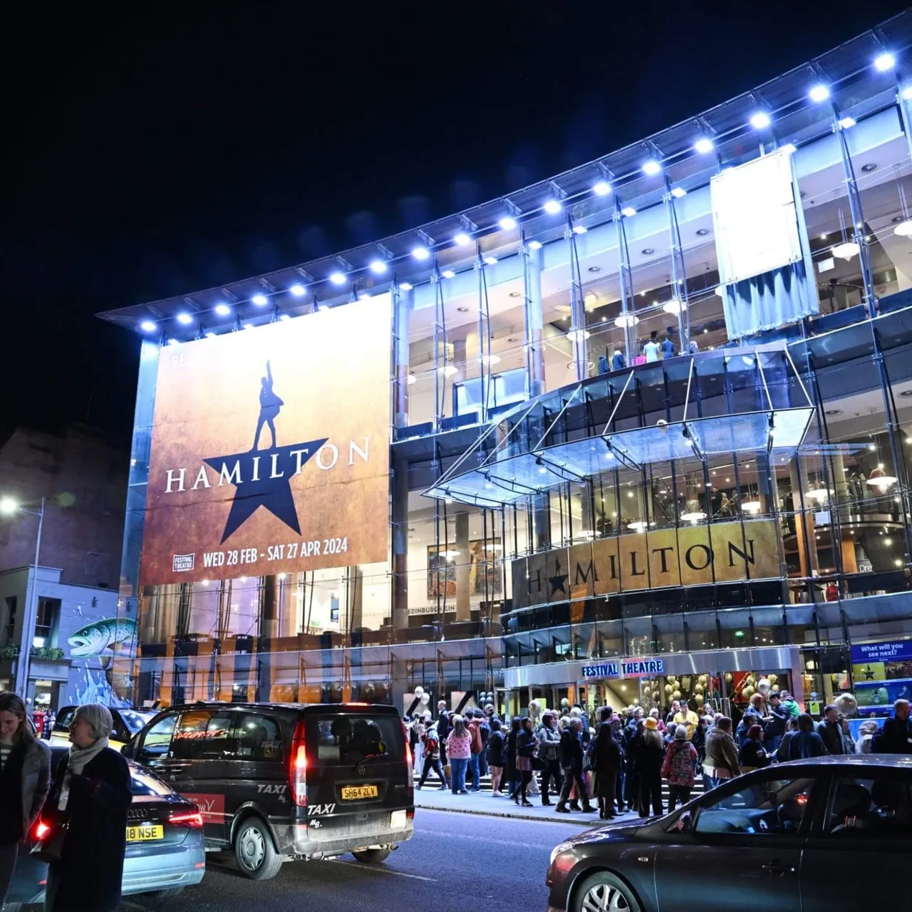 Illuminated theatre building at night with Hamilton musical advertisement, crowds of people outside, and taxis parked in front.