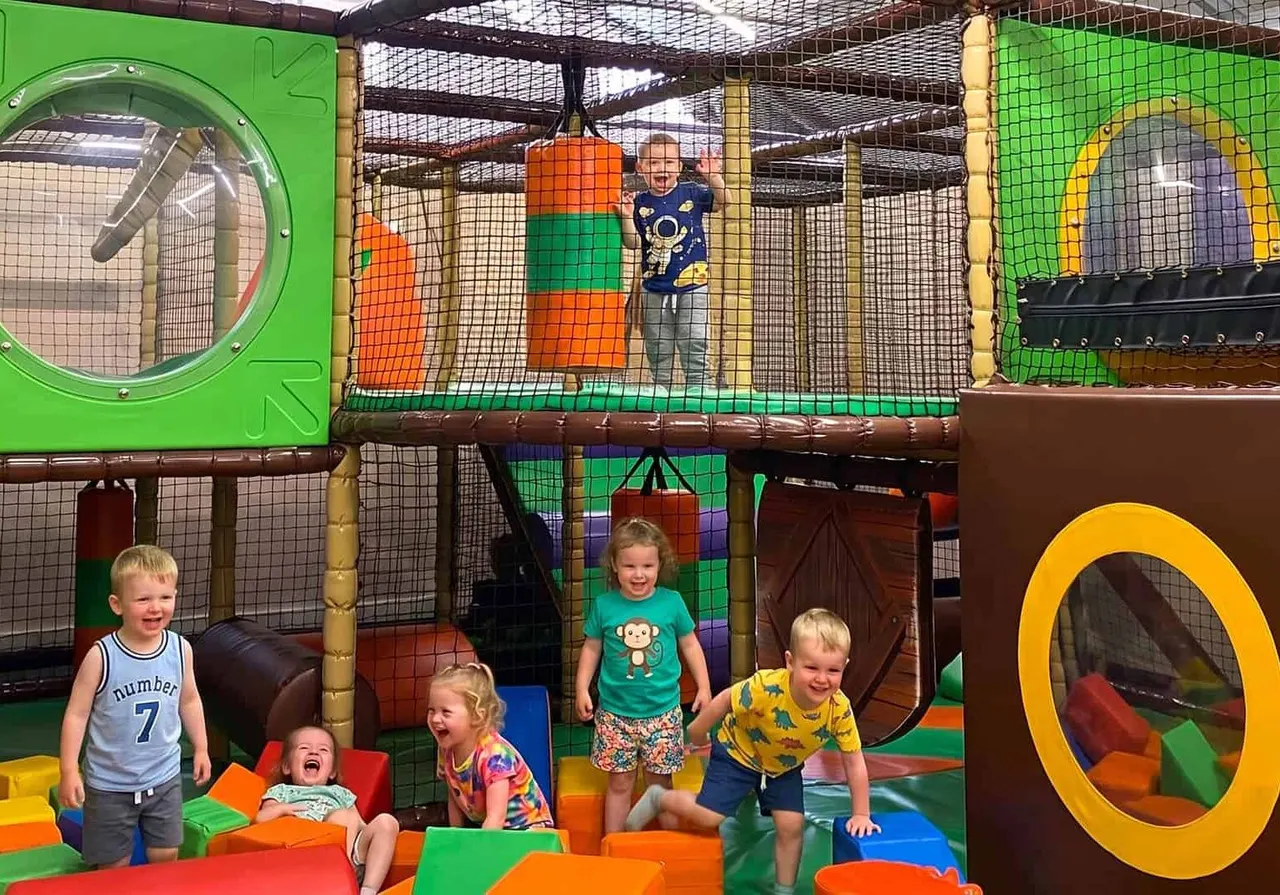 Young children playing and climbing in a colourful indoor soft play area with various foam structures and netting.