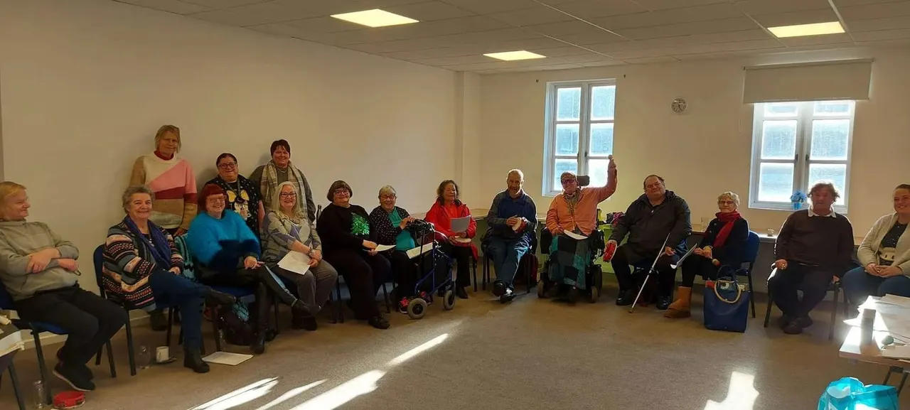 Group of older adults seated in a circle in a bright community hall during a singing or social activity session.