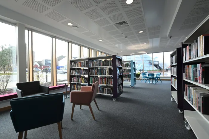 Modern library interior with armchairs, bookshelves and large windows overlooking a car park and mountains.