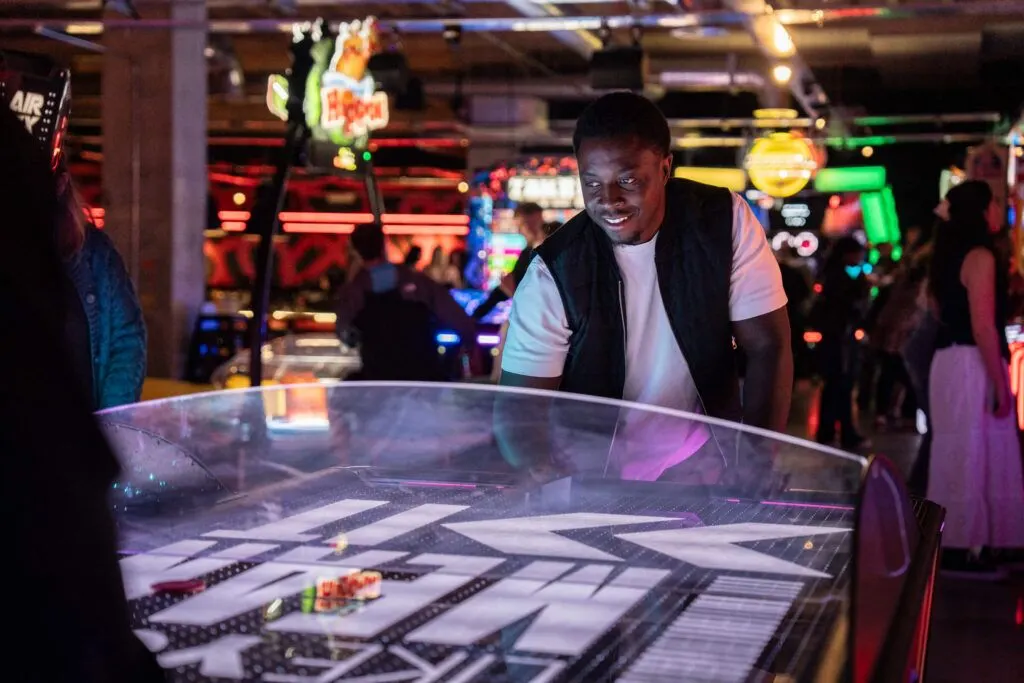 Man playing air hockey at a neon-lit indoor entertainment venue with arcade games and other guests in the background.
