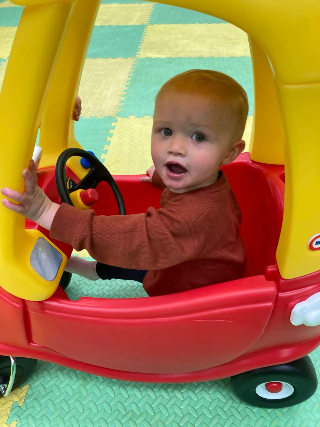 Toddler in red seat holding steering wheel of toy ride-on car indoors.