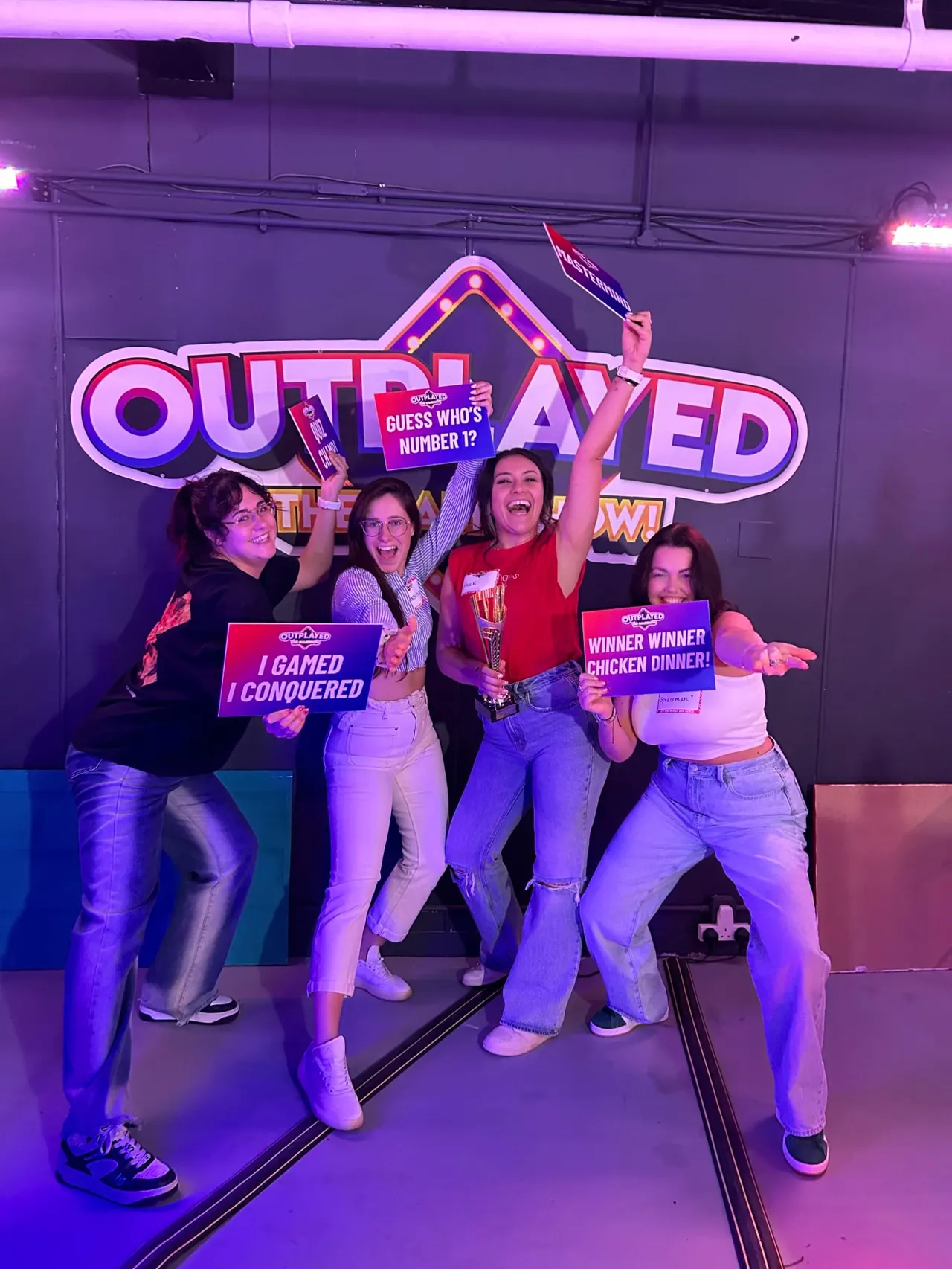 Four women posing playfully in front of a neon-lit arcade game sign, holding gaming prize cards.
