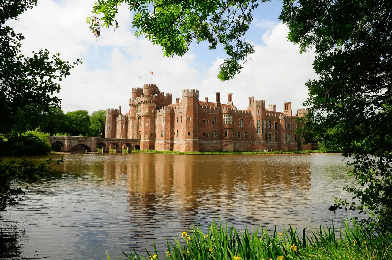 Red brick castle with crenellated towers reflected in a moat, surrounded by trees and green vegetation.