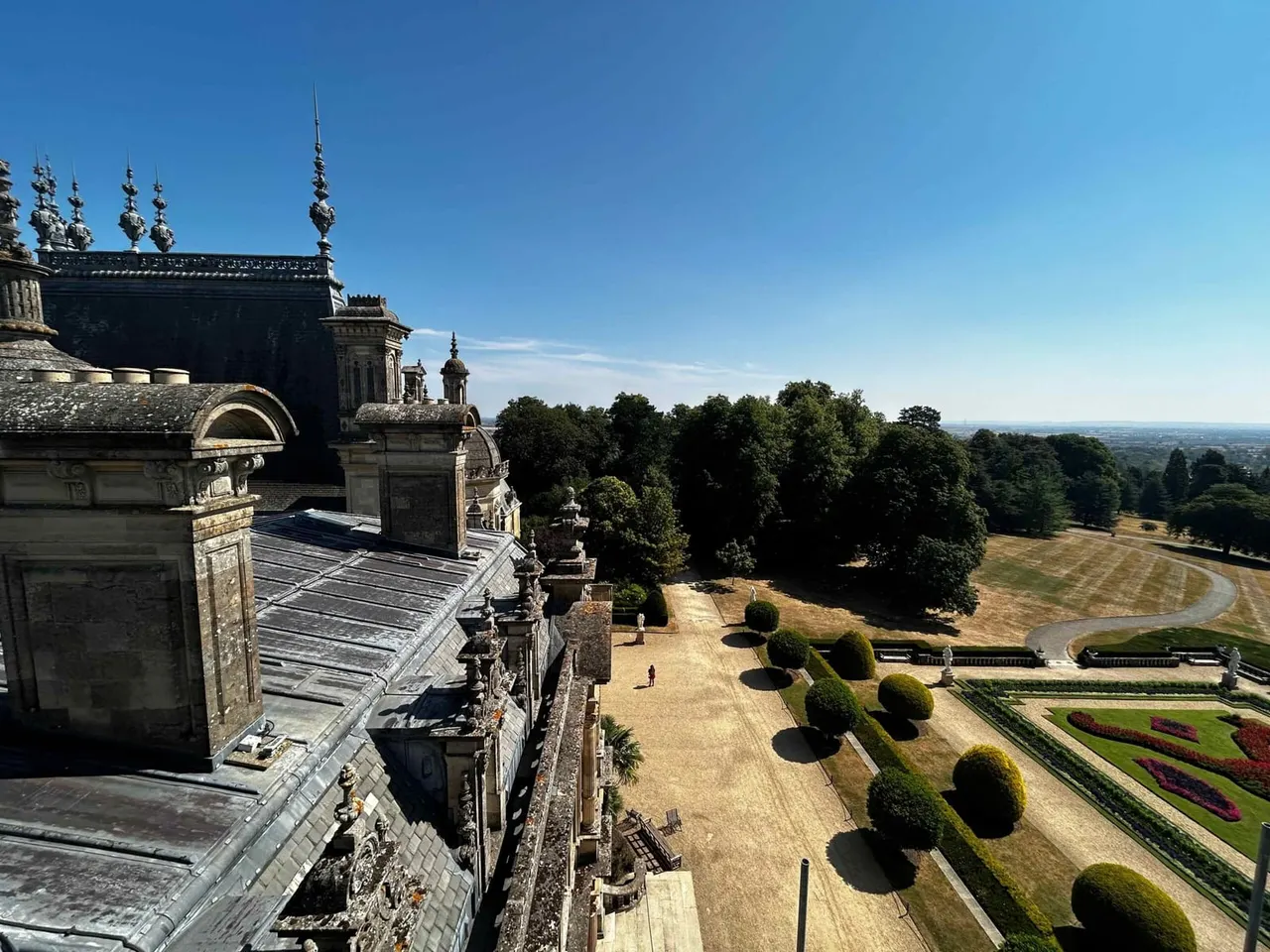 Ornate stone mansion with spires overlooks formal gardens, manicured hedges and parkland under clear blue sky.