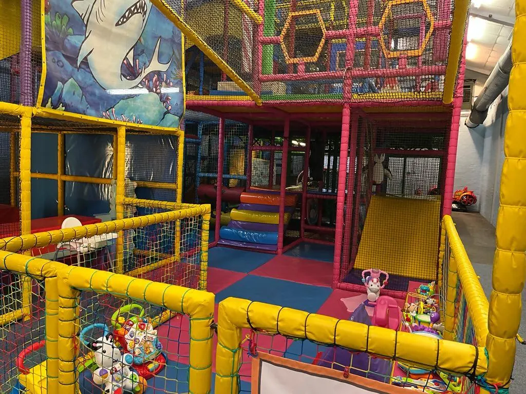 Colourful indoor soft play area with yellow-framed structures, red and pink padded sections, ball pits, slides and climbing equipment for young children.