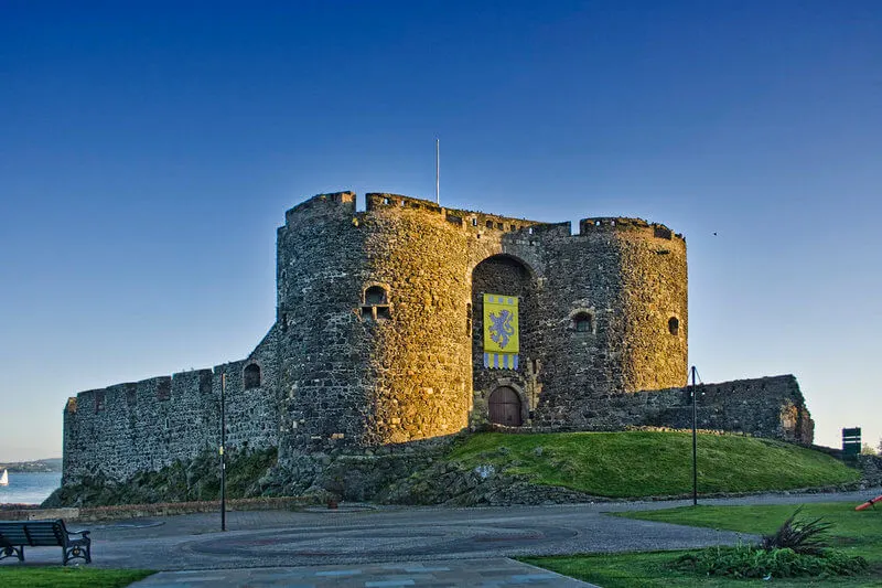 Historic stone fortress tower with crenellated walls on grassy mound beside viewing area under clear blue sky.