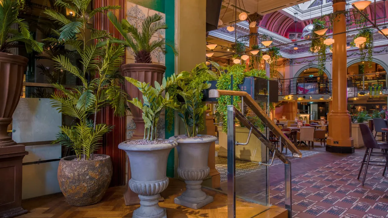 Grand interior of a hotel or restaurant atrium with potted plants, decorative urns, elegant lighting, and multiple levels visible above.