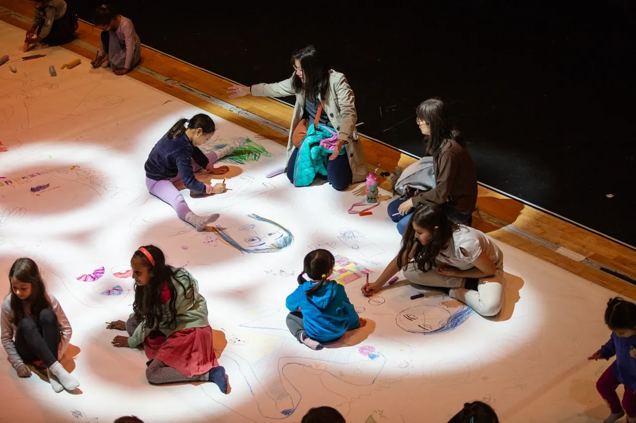 Children and adults drawing together on a large illuminated canvas floor during an interactive art activity.