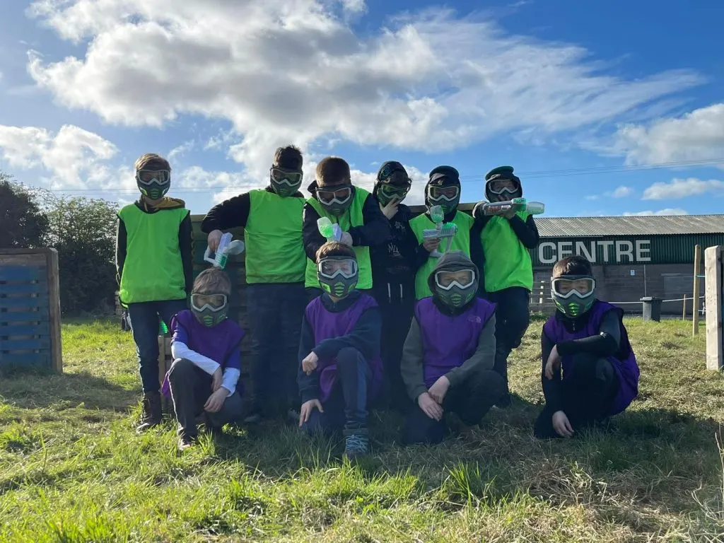 A group of young people wearing protective goggles and coloured tabards pose together outdoors at a paintball facility.
