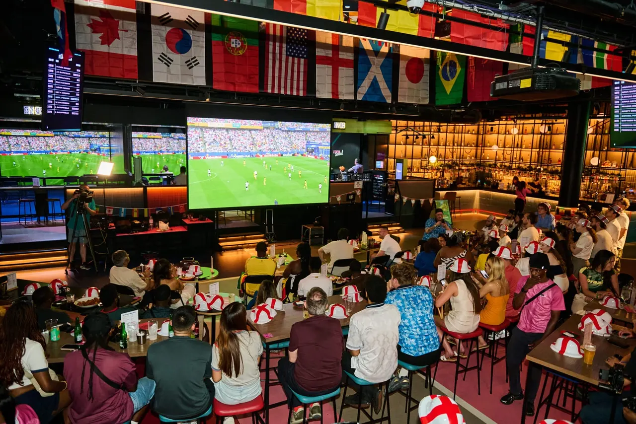 Crowds of spectators watch a football match on large screens whilst seated at tables in a sports bar venue decorated with international flags.