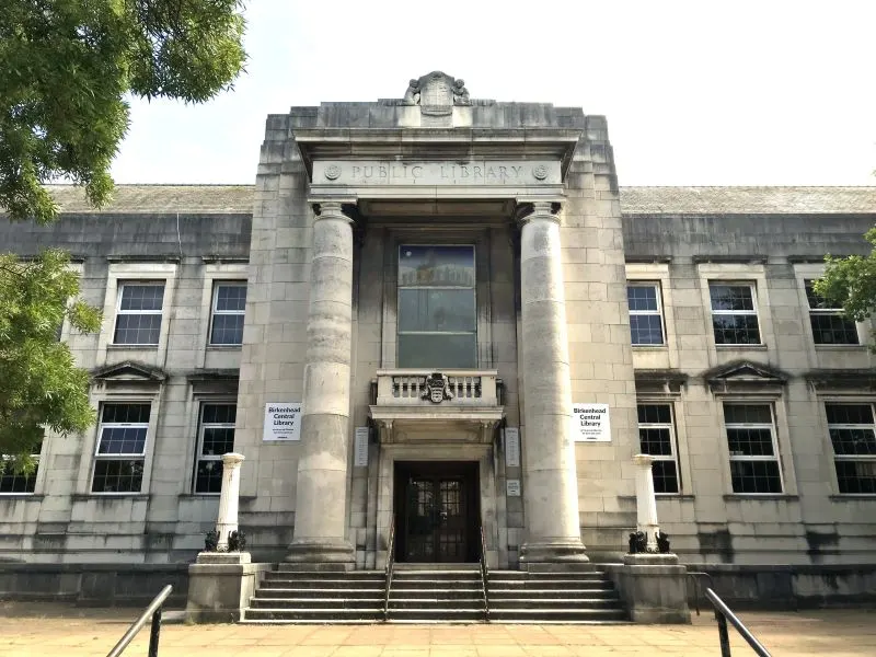 Historic stone public library building with classical columns and entrance steps, framed by green foliage.