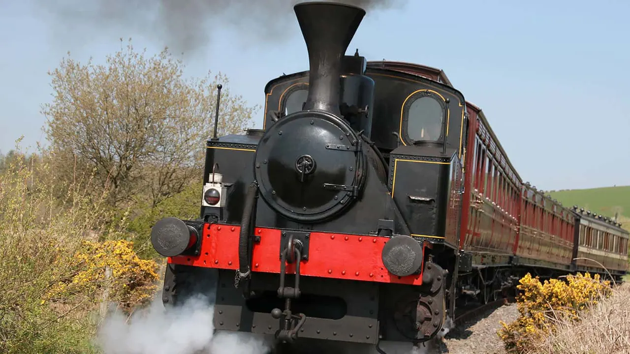 Heritage steam locomotive with red and black livery pulling vintage passenger carriages through countryside.