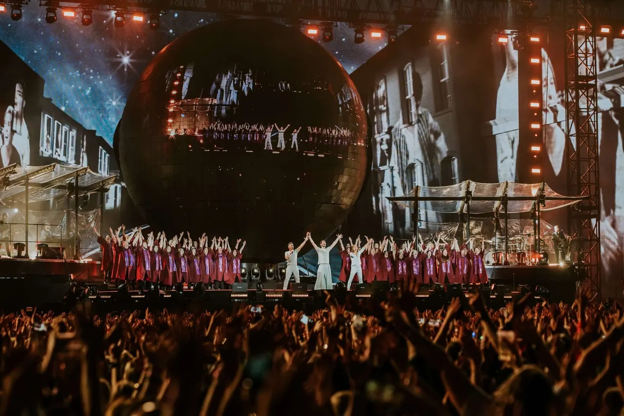 Large crowd with raised hands watching performers on an illuminated stage with choir members in burgundy robes and giant spherical set piece.