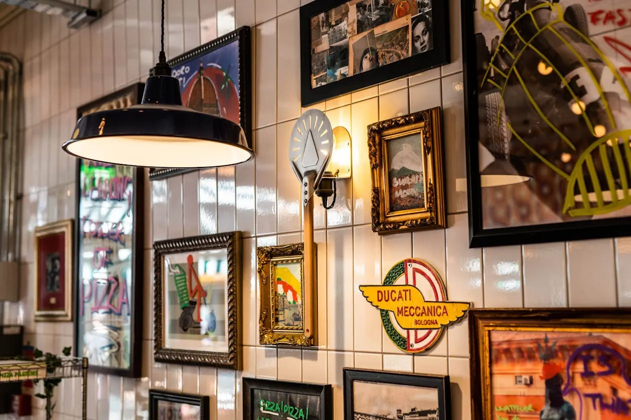 Interior wall of Italian restaurant decorated with vintage framed photographs, neon signs, and a hanging black pendant light.