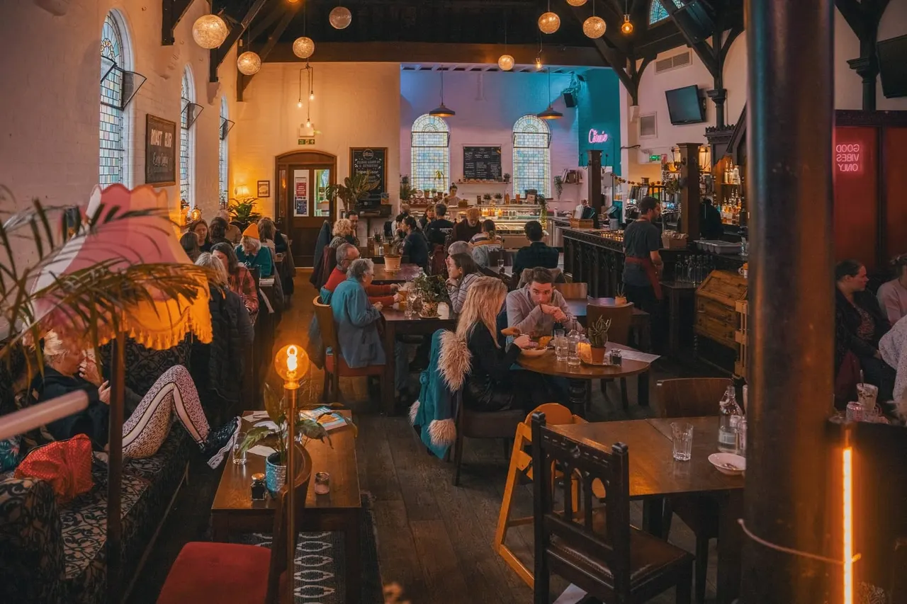 A busy restaurant interior with diners seated at tables, a bar counter, pendant lighting, and warm golden and blue ambient lighting throughout the space.