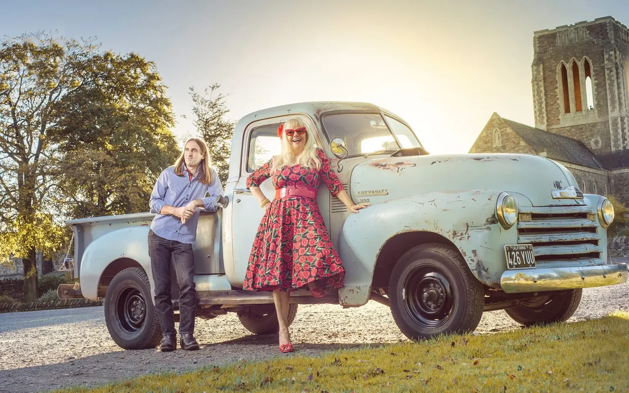 Two people pose with a vintage light blue pickup truck beside a country church at sunset.