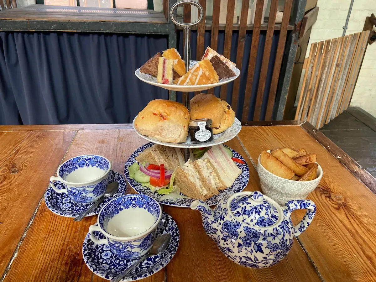 Tiered afternoon tea stand with sandwiches, scones, pastries, and cakes, alongside teapot and cups on wooden table.