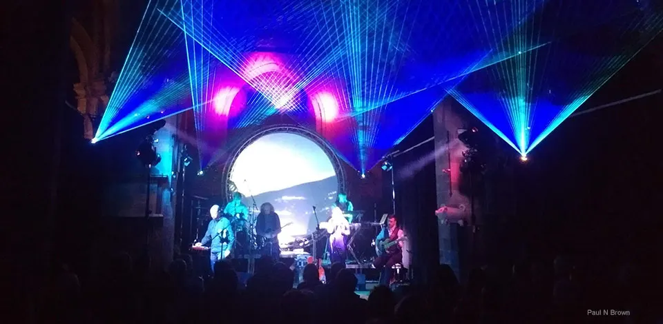 Live band performs on stage beneath dramatic blue and pink laser light display with audience silhouettes.