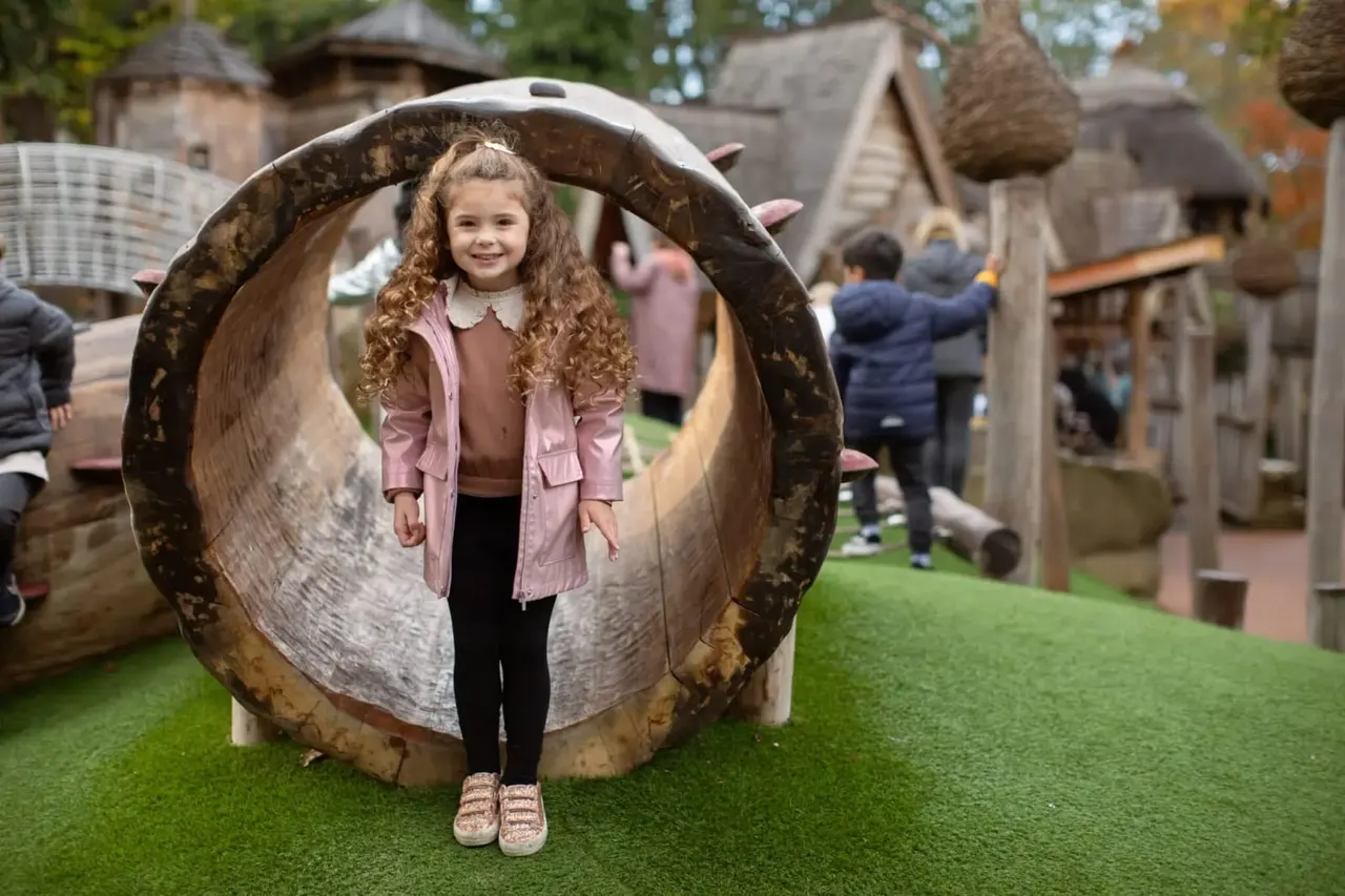 Young girl with curly hair standing inside a large hollow log tunnel at an outdoor play area, with stone buildings and other children in the background.