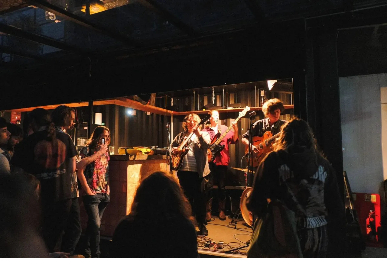 A live band performs on an indoor stage with guitars and drums whilst an audience watches from the darkened venue floor.