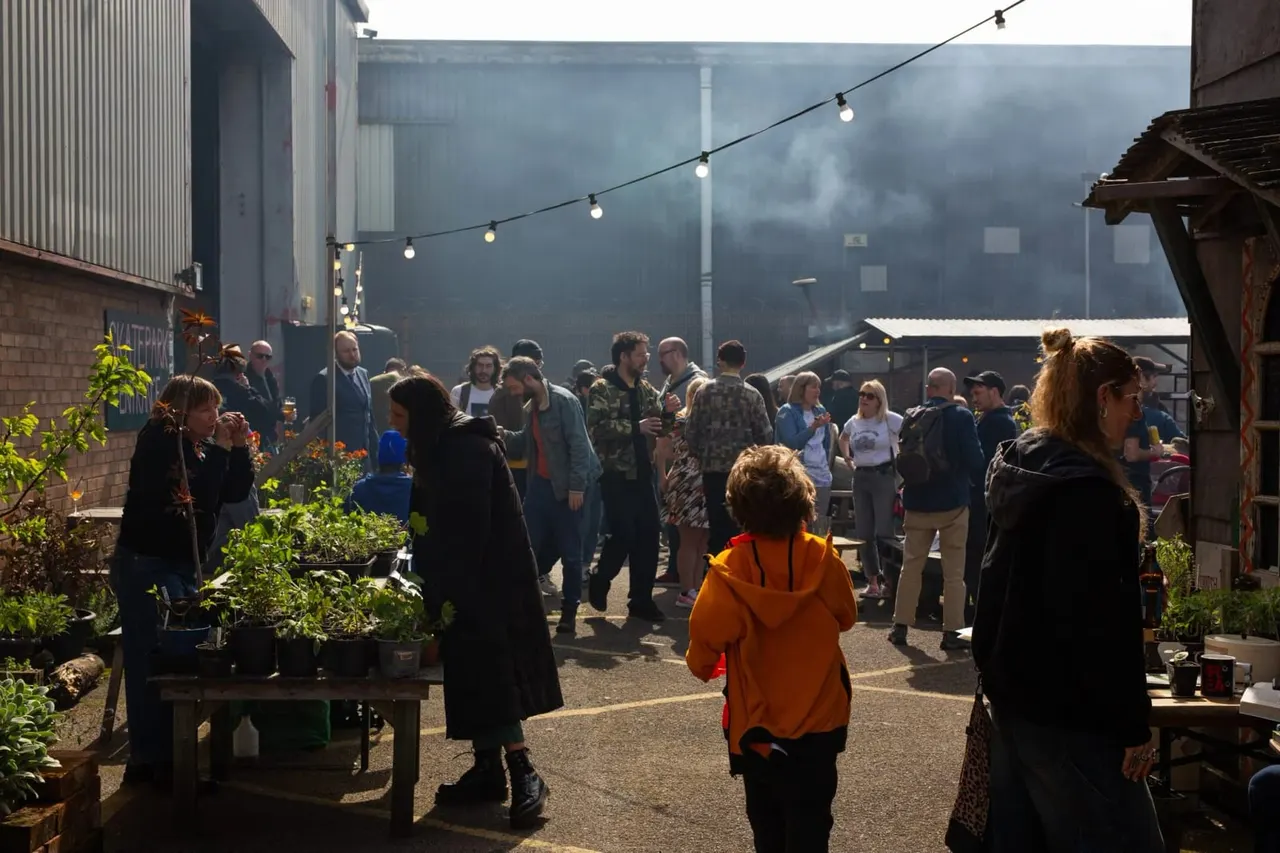 Large crowd of visitors mingling in an industrial courtyard with string lights, potted plants, and corrugated metal buildings.