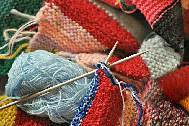 Knitting needles with blue yarn and colourful knitted fabric swatches in progress.