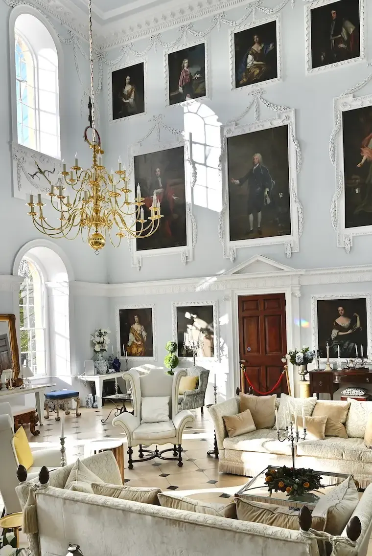Ornate white reception room with period furniture, gold chandelier, arched windows and portrait paintings on walls.