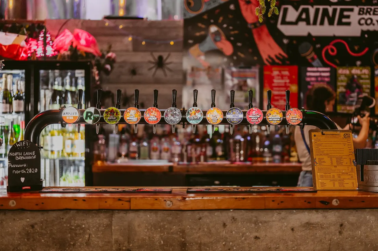 A row of colourful beer pump taps at a bar counter, with festive decorations and spirits bottles visible in the blurred background.