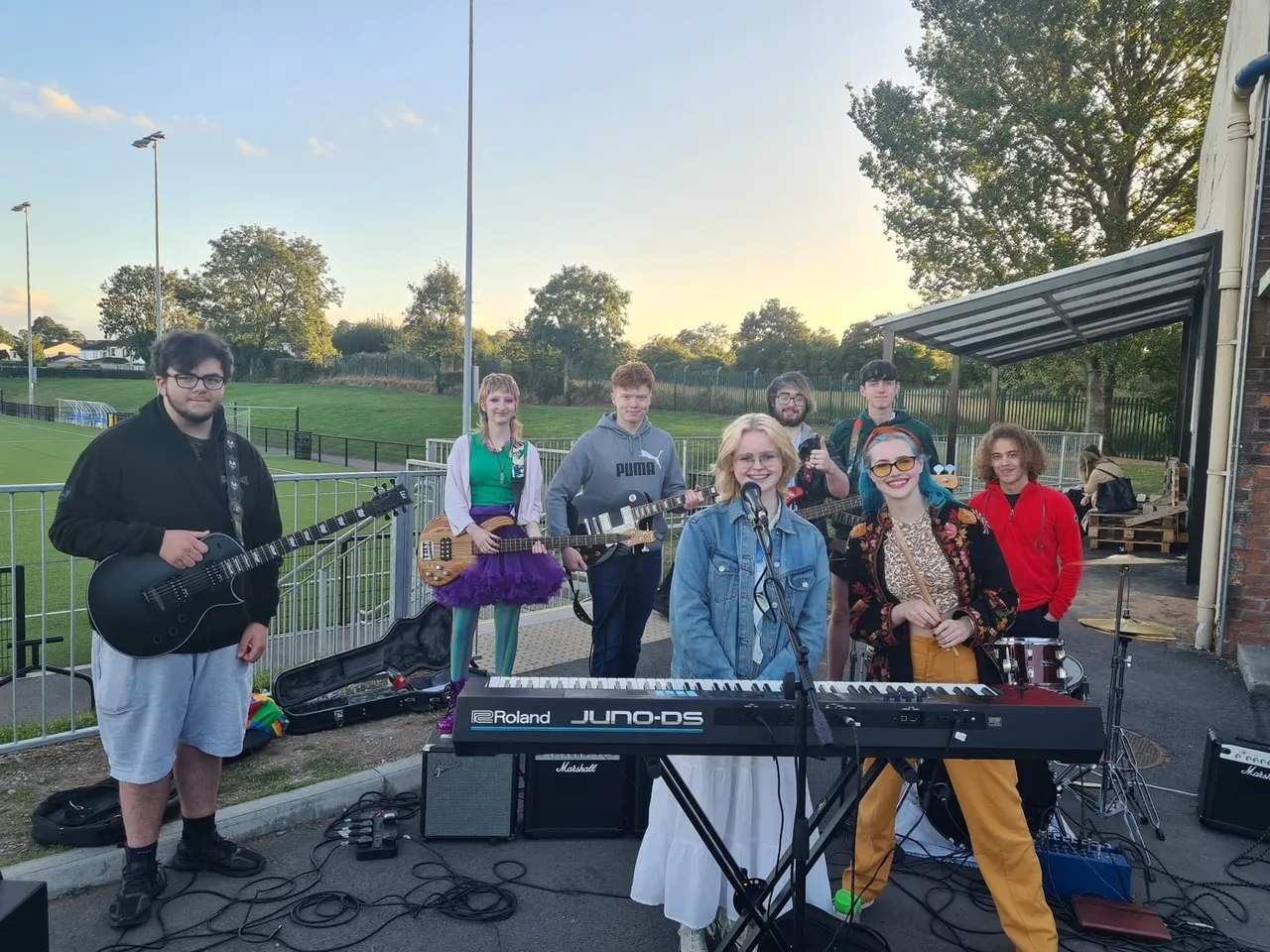 A band of young musicians performing outdoors at a sports field venue, with a singer at a keyboard, guitarists, and drummer under a covered shelter.