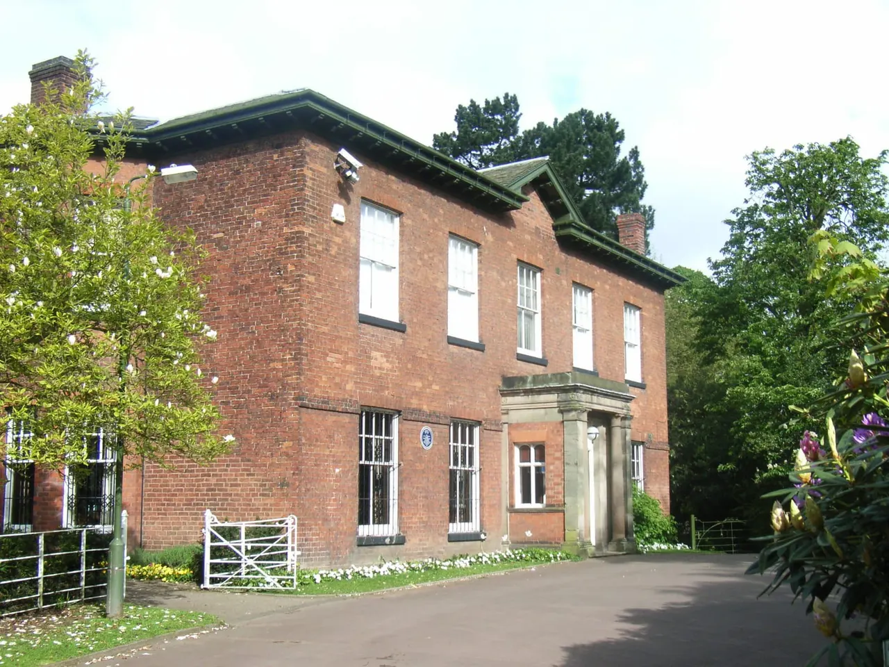 Victorian red brick house with green roof and stone entrance portico, surrounded by mature trees and flowering plants.