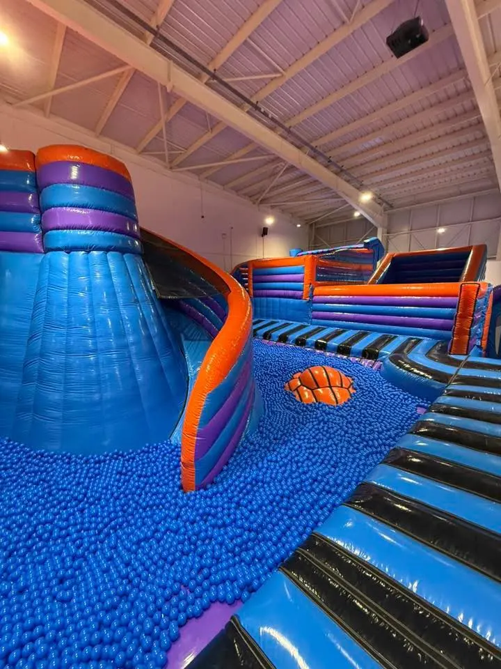 Indoor play area with colourful inflatable structures including slides and climbing obstacles on a blue foam floor.