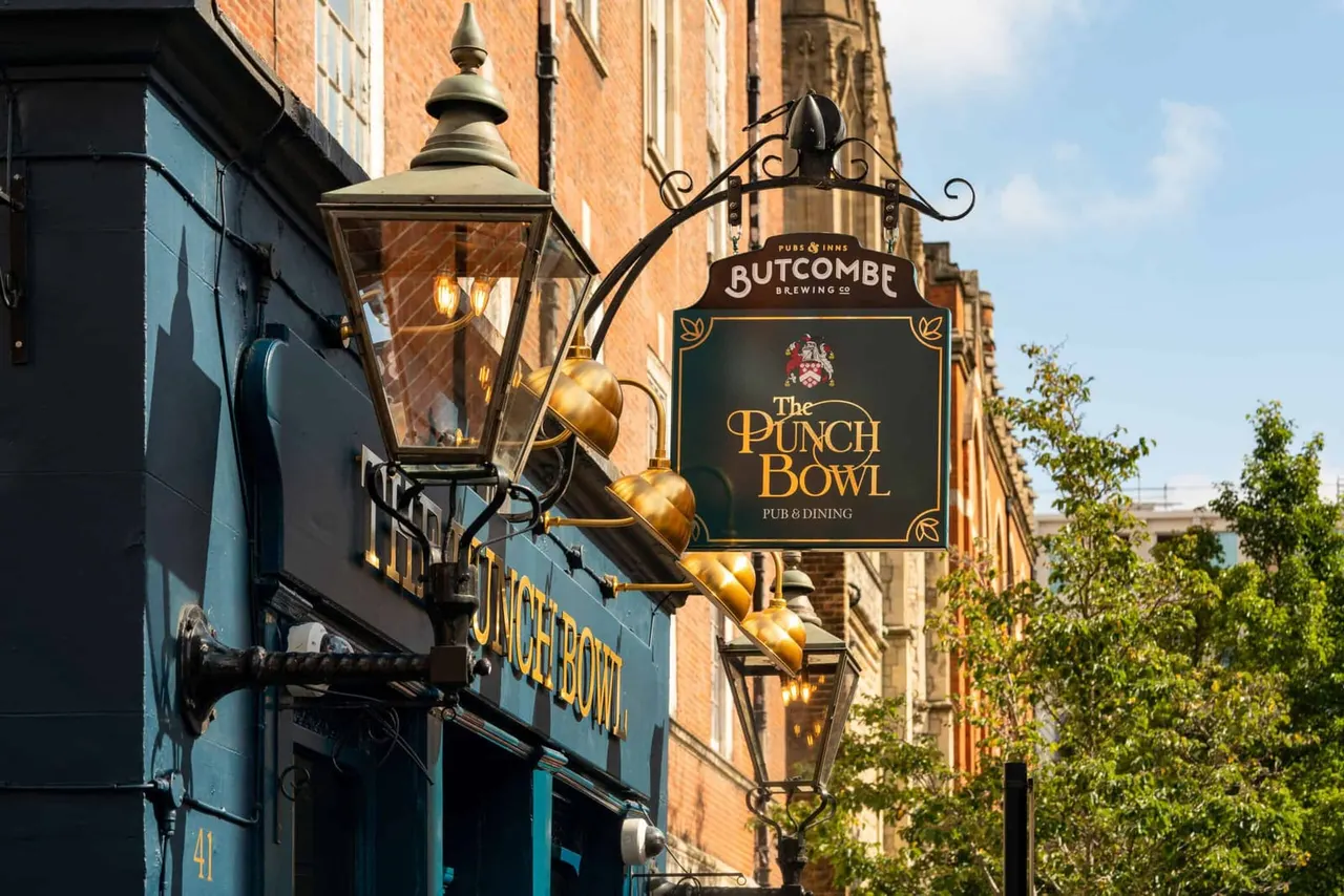 Ornate brass hanging pub signs for The Punch Bowl and Butcombe Brewing Co on a red brick building exterior, with decorative street lamps.