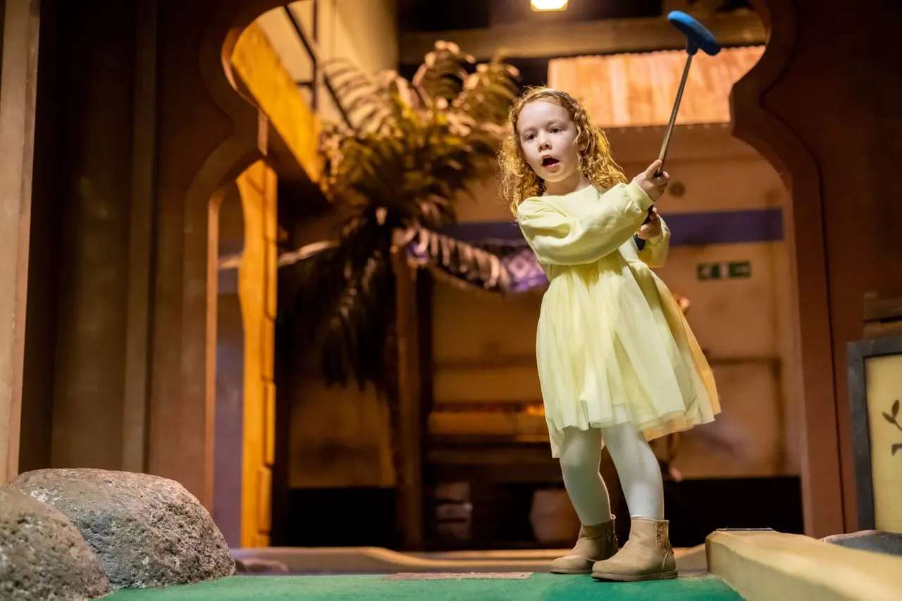 Child in pale yellow dress playing mini golf, holding putter with surprised expression.