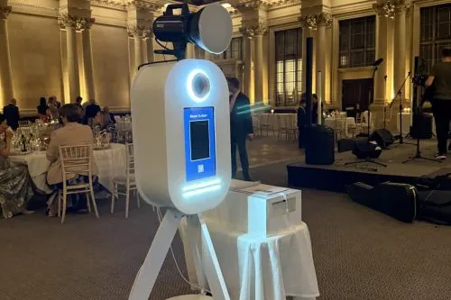 A white robotic photo booth with blue illuminated accents stands in an ornate ballroom during an event, with guests and dining tables visible in the background.