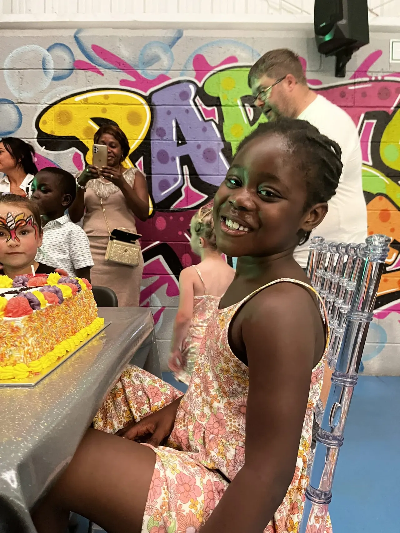 Children and adults celebrating at a party with a decorated cake and colourful graffiti wall backdrop.