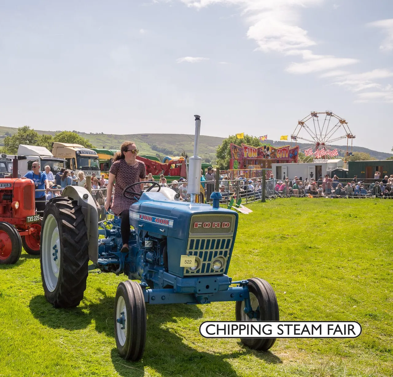 A woman stands on a blue vintage Ford tractor at an outdoor steam fair, with a funfair and crowds visible behind.
