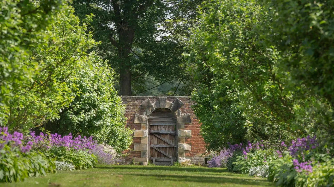 A brick archway with a wooden door seen through lush green hedges and purple flowering plants in a garden setting.