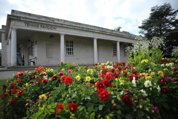 A white classical pavilion building with columned portico, with vibrant red, yellow and white flowers in beds in the foreground.
