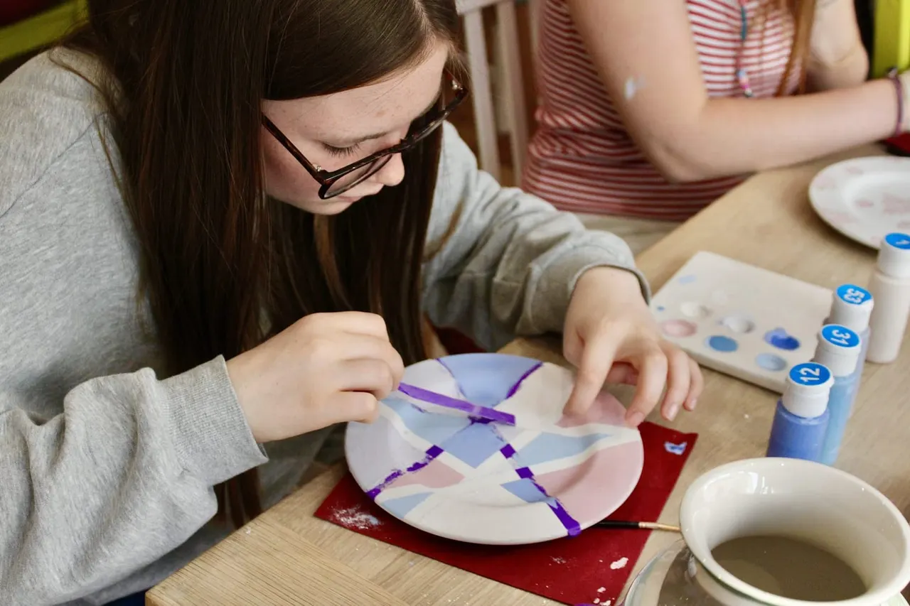 Child wearing glasses painting a ceramic plate with purple paint at a pottery painting table.