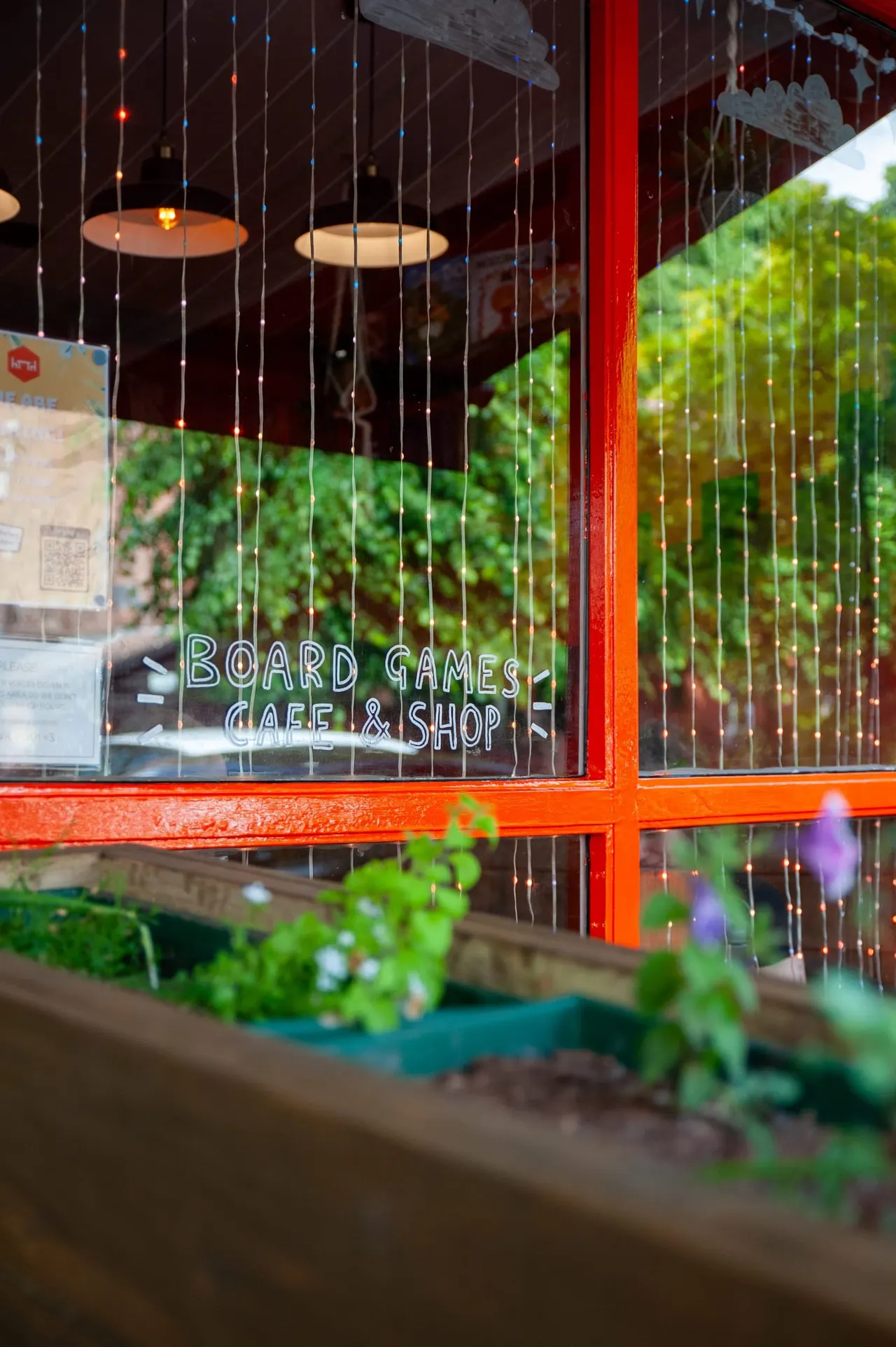 Board games café storefront with red window frame, hanging pendant lights and beaded curtains, viewed from outside with plants in foreground.