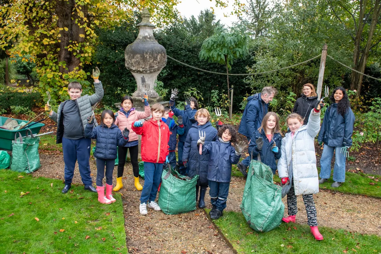 Group of children and adults holding garden tools and waste bags during a garden activity in an autumnal woodland setting.