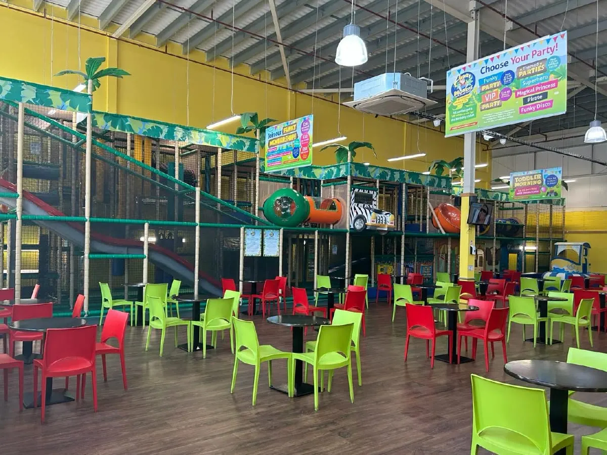 Colourful indoor play area with bright red and lime green chairs around tables, climbing frames and soft play equipment visible behind netting.