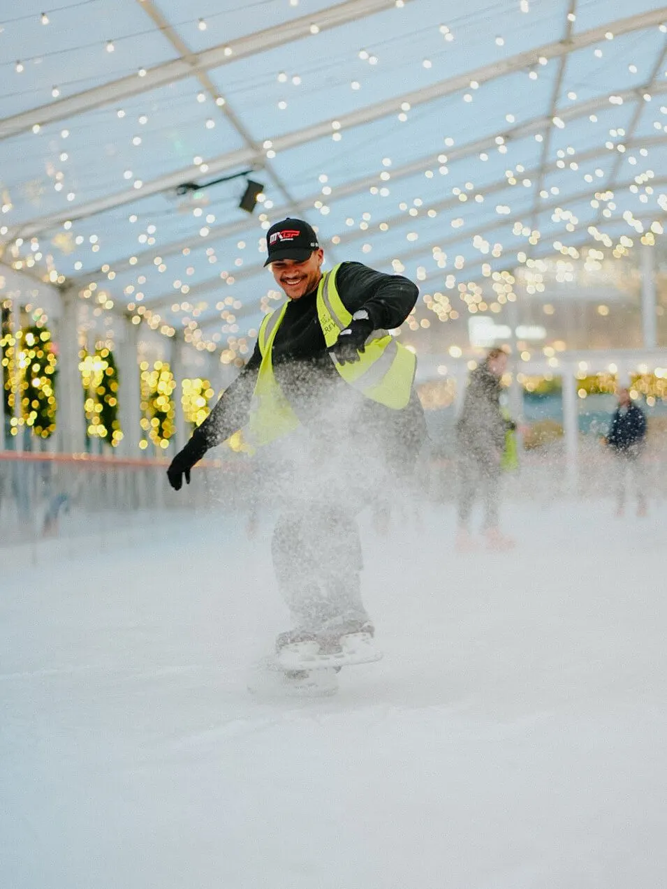 Person ice skating indoors under string lights, wearing a high-visibility jacket and cap.